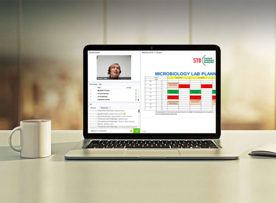 A laptop open on a modern office desk next to a white ceramic mug, displaying an online technical webinar interface with a slide titled 'MICROBIOLOGY LAB PLANNING' and a live video feed of speaker Lidia Lianiuka Lenivko.