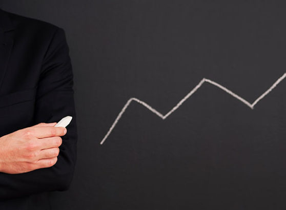 Midsection of a business consultant in a dark suit and patterned blue tie holding a piece of chalk, standing next to an upward-trending zigzag growth line drawn on a blackboard.