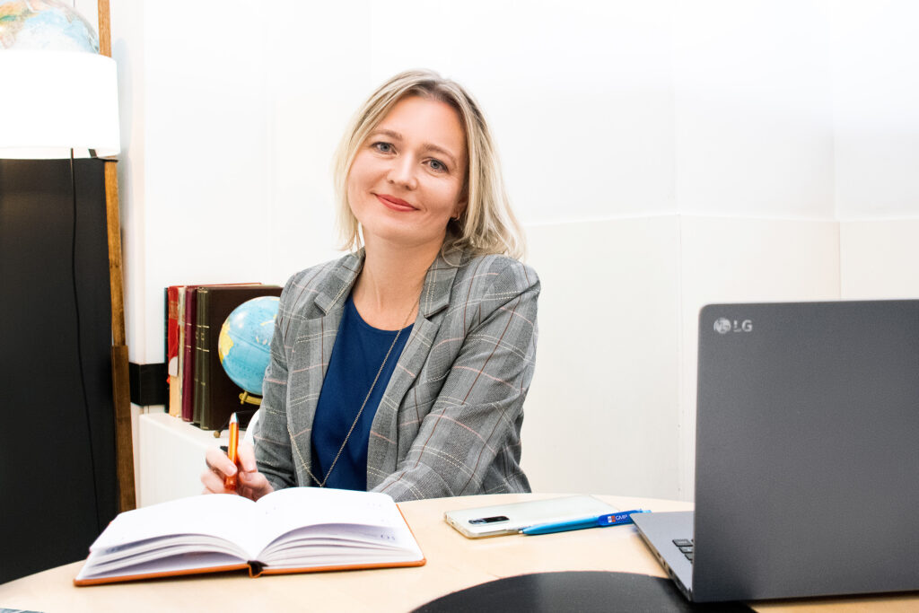 A professional portrait of Lidia Lianiuka Lenivko, founder of RuGMP, smiling in a grey plaid blazer and blue blouse at her desk, with a desktop globe and books visible in the background.