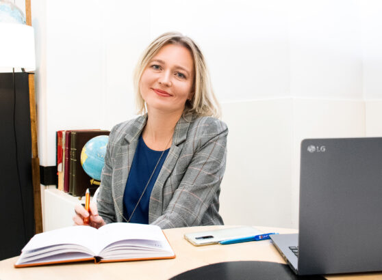 A professional portrait of Lidia Lianiuka Lenivko, founder of RuGMP, smiling in a grey plaid blazer and blue blouse at her desk, with a desktop globe and books visible in the background.