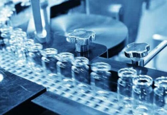 Close-up of clear glass pharmaceutical vials moving rapidly along an automated conveyor belt track in a manufacturing plant under bright blue technical lighting.