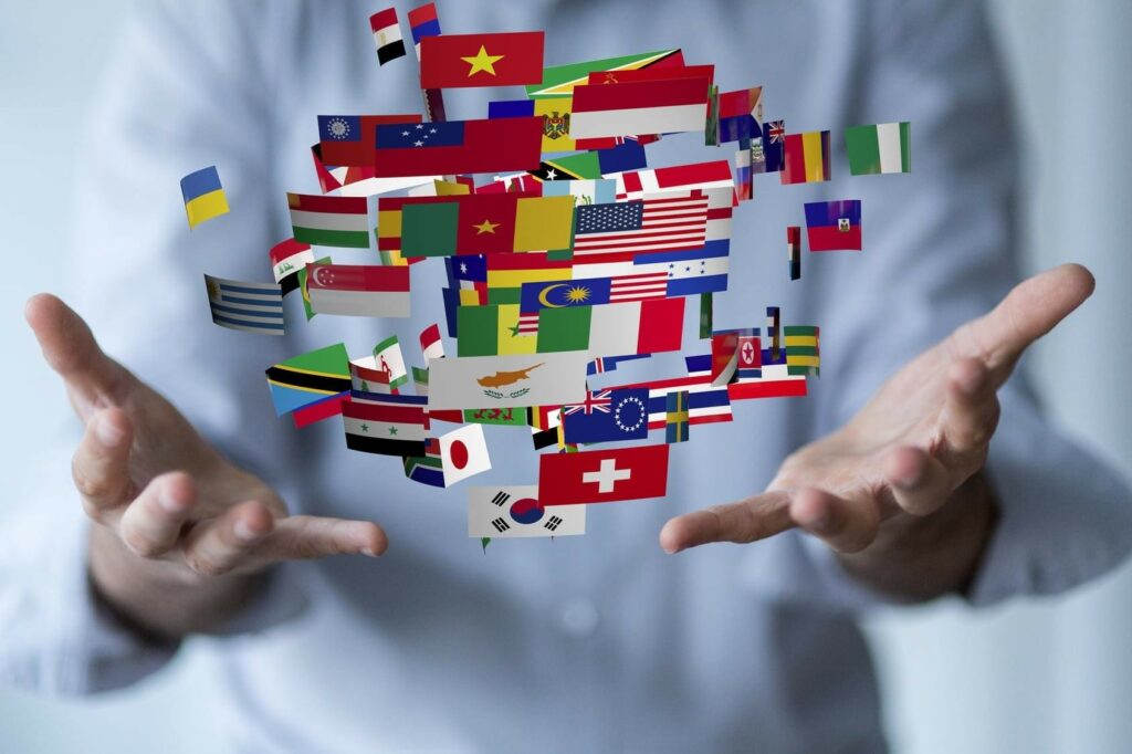 Primer plano de las manos abiertas de un profesional sosteniendo de manera simbólica una nube flotante y dinámica de múltiples banderas nacionales de diferentes países del mundo (como Vietnam, Estados Unidos, Suiza, Ucrania y Corea del Sur), representando los servicios globales de traducción e interpretación lingüística.