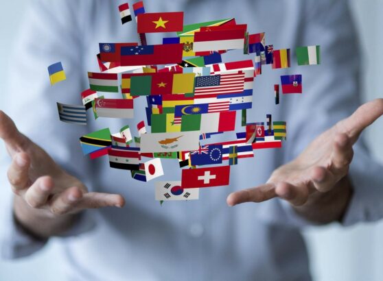 Primer plano de las manos abiertas de un profesional sosteniendo de manera simbólica una nube flotante y dinámica de múltiples banderas nacionales de diferentes países del mundo (como Vietnam, Estados Unidos, Suiza, Ucrania y Corea del Sur), representando los servicios globales de traducción e interpretación lingüística.