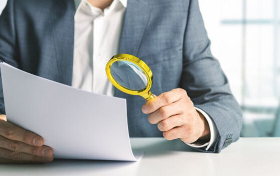 Close-up of a professional in a gray suit sitting at a white desk, holding a golden magnifying glass to closely scrutinize a white document in a bright office environment.